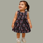 Black sleeveless slub cotton frock for baby girls with red floral block prints and a pleated flare, displayed flat on a white fur background with floral basket hanger.