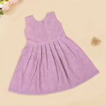 Mauve Hakoba frock for baby girls, sleeveless and pleated with soft embroidery, laid flat on a beige background beside floral accents and a heart-shaped wicker prop.