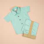 Teal blue silk dhoti shirt set for baby boys featuring a wooden-button shirt and gold-bordered dhoti, displayed flat on a beige background with floral props.