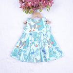 Girls' sleeveless frock in pastel blue cotton with butterfly and floral print, displayed with a wooden hanger under pink orchids – Boon Babies summer casual wear collection.