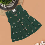Floral green silk sleeveless frock for baby girls with wooden buttons and subtle motifs, displayed on a beige backdrop with soft floral accents and woven mat.