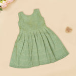Pastel green sleeveless Hakoba frock for baby girls, featuring floral embroidery and pleated skirt design. Lightweight cotton fabric, displayed flat against a soft beige background with a decorative heart and flowers.