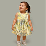 Yellow sleeveless floral chiffon cotton frock for baby girls, with pleated skirt and pastel tones, displayed on a wooden hanger over a white fur background and floral décor.