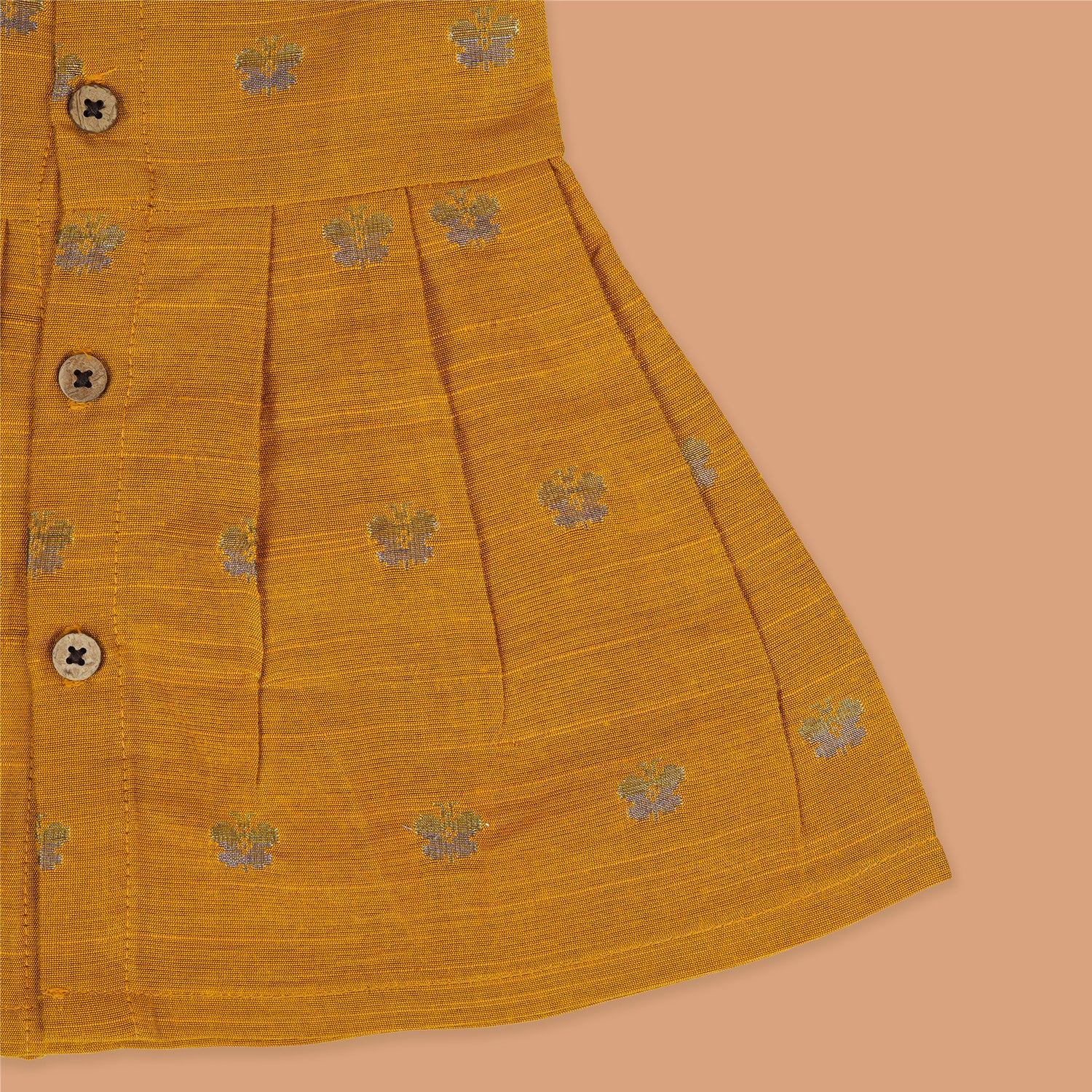 Yellow silk sleeveless ethnic frock for baby girls with floral woven motifs and wooden button closure, displayed on a peach background with floral and natural props.