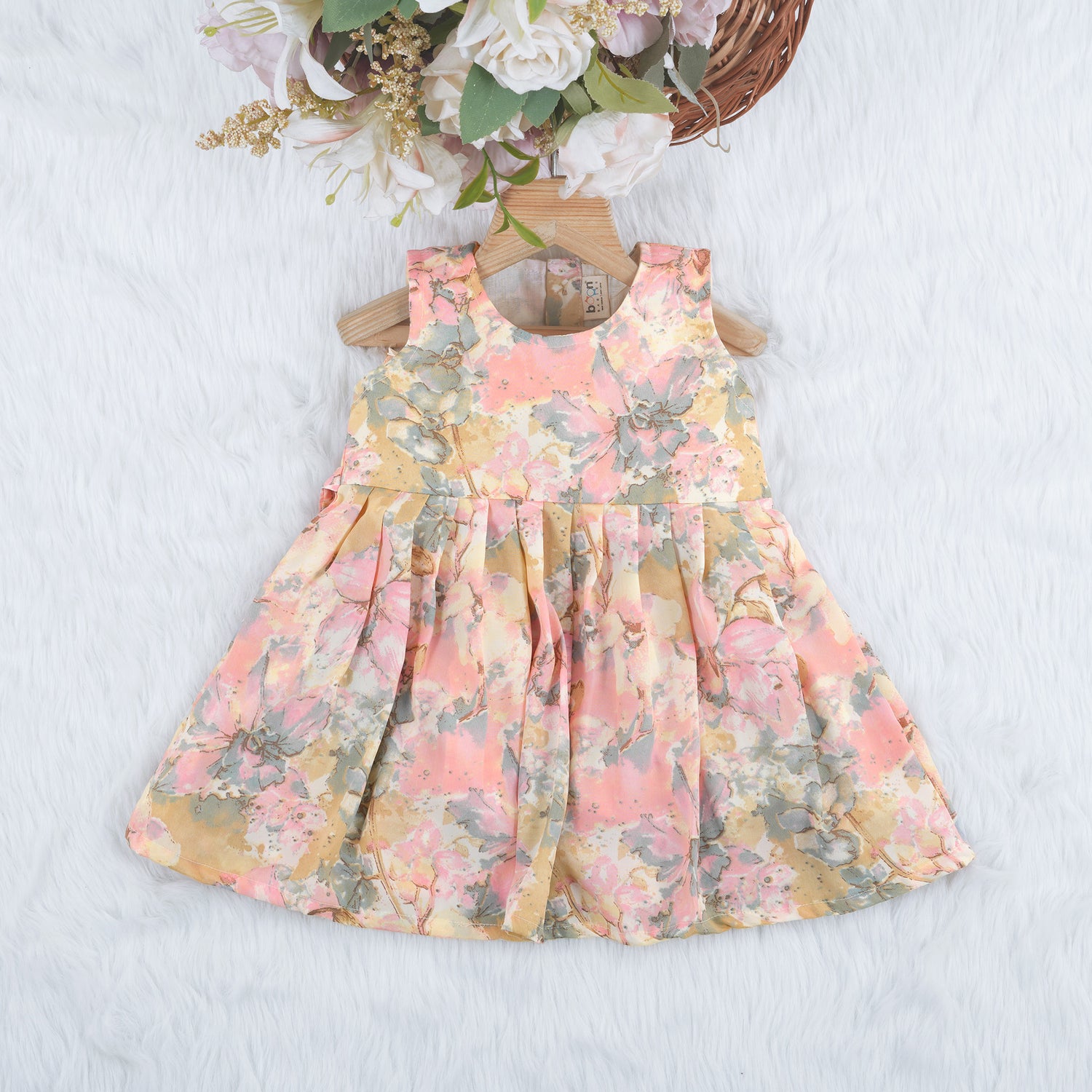Floral printed sleeveless frock for baby girls in pastel shades of pink, beige, and grey, made from chiffon cotton and displayed on a wooden hanger over white fur with flower props.