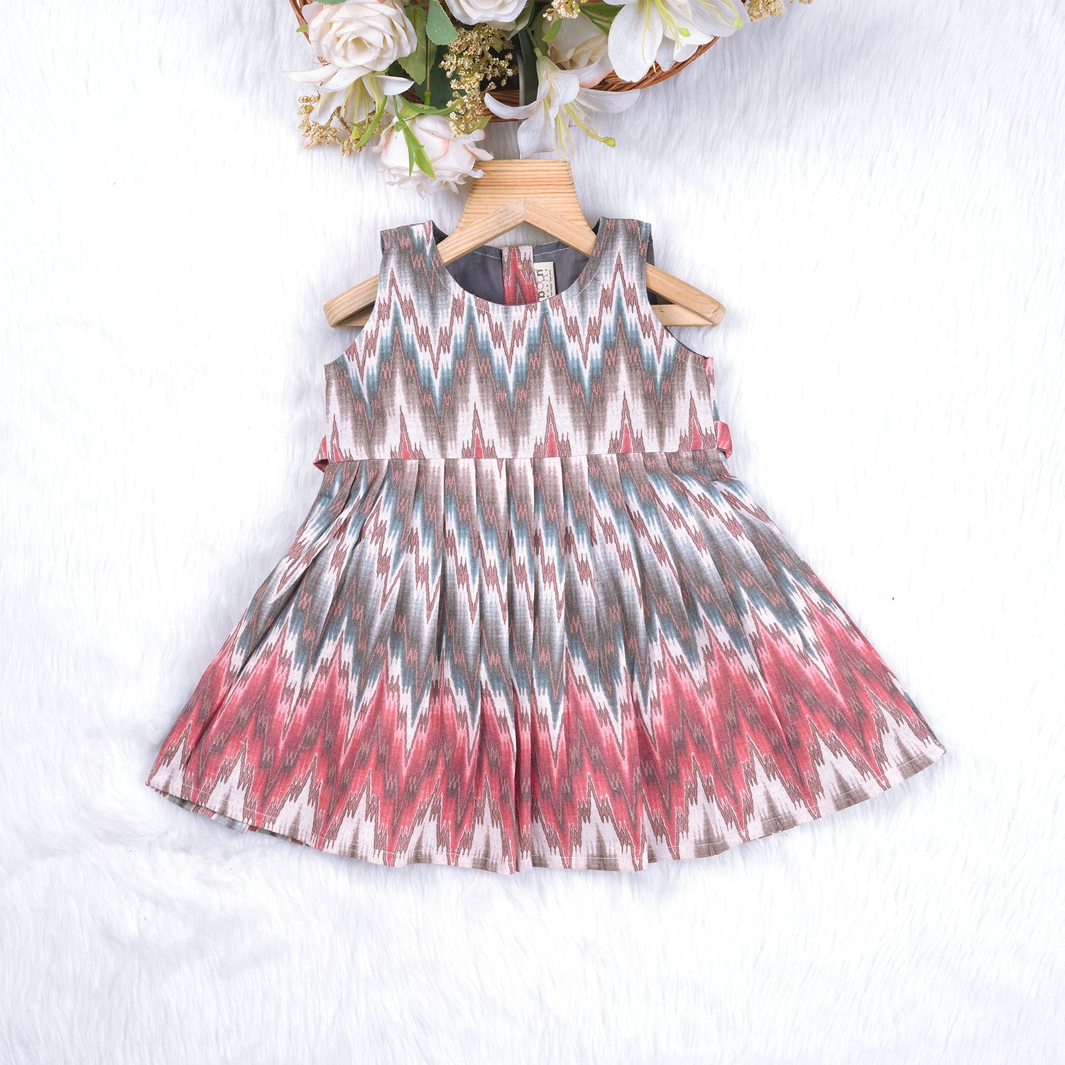 Girls’ sleeveless frock in multicolor Ikat print, made from cotton – displayed flat on white fur background with floral props – Boon Babies casual summer wear for girls aged 12M to 5Y.
