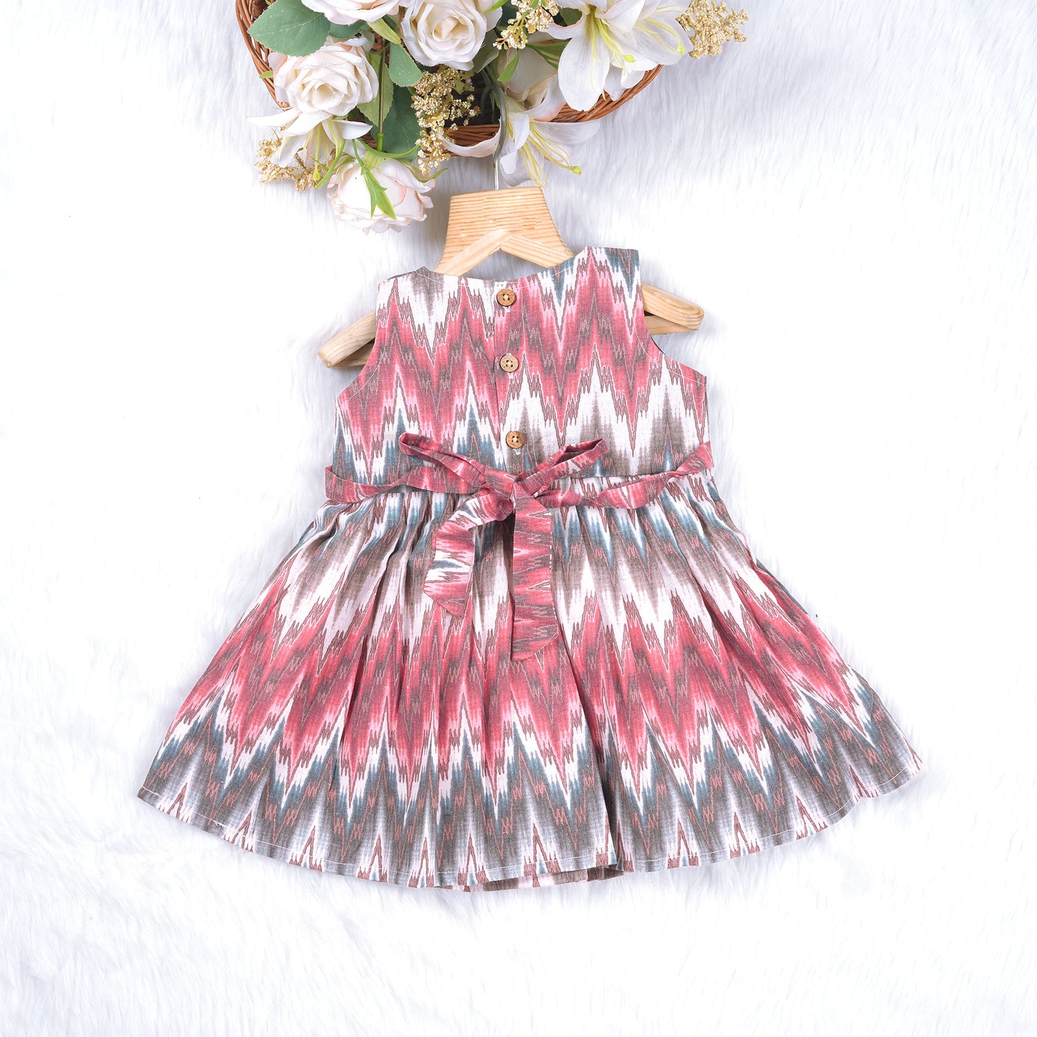 Girls’ sleeveless frock in multicolor Ikat print, made from cotton – displayed flat on white fur background with floral props – Boon Babies casual summer wear for girls aged 12M to 5Y.
