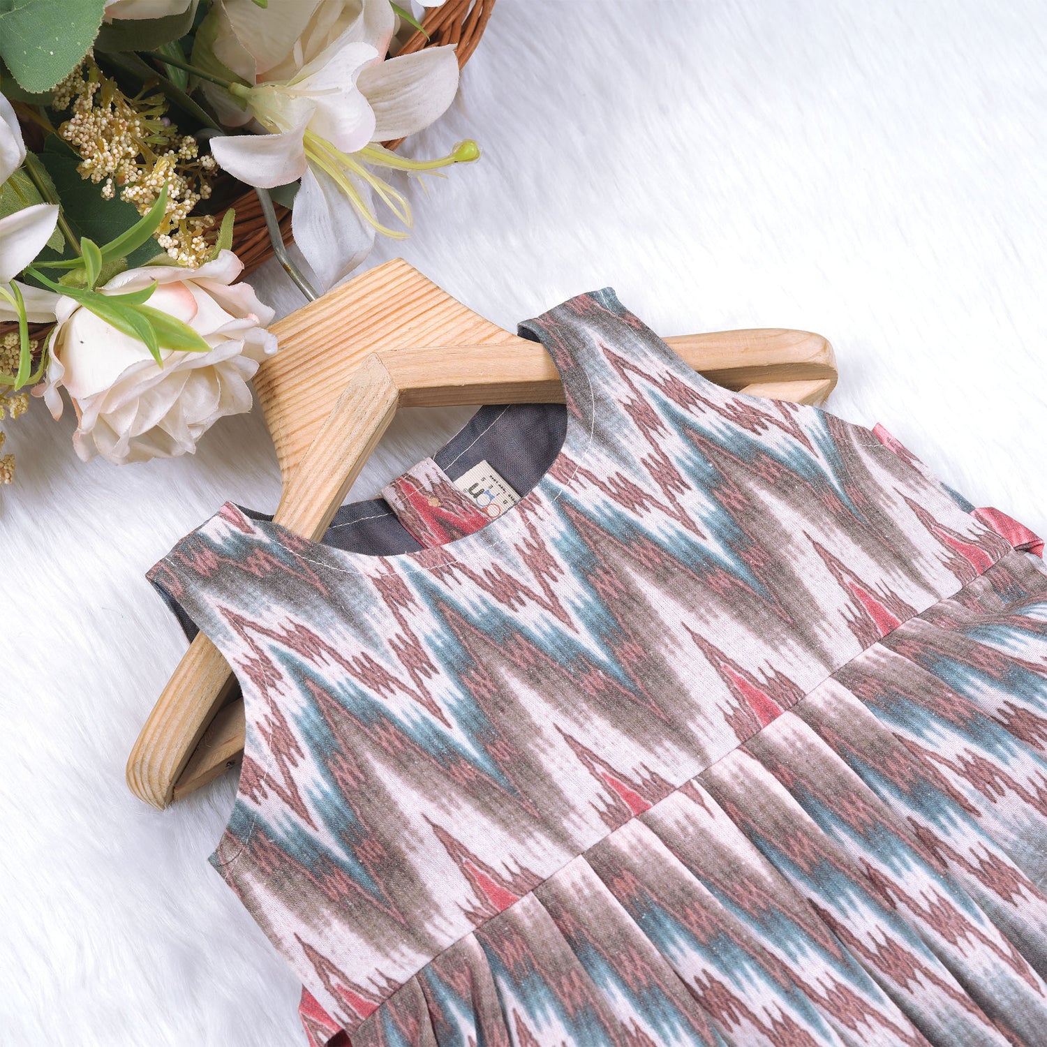 Girls’ sleeveless frock in multicolor Ikat print, made from cotton – displayed flat on white fur background with floral props – Boon Babies casual summer wear for girls aged 12M to 5Y.
