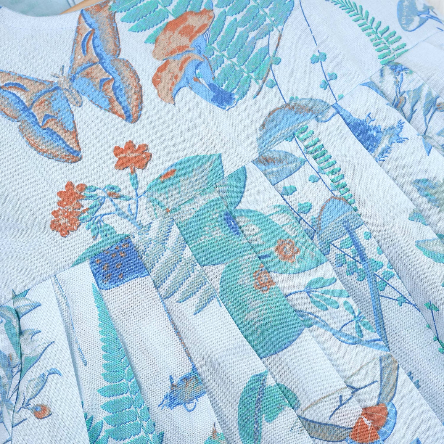 Girls' sleeveless frock in pastel blue cotton with butterfly and floral print, displayed with a wooden hanger under pink orchids – Boon Babies summer casual wear collection.
