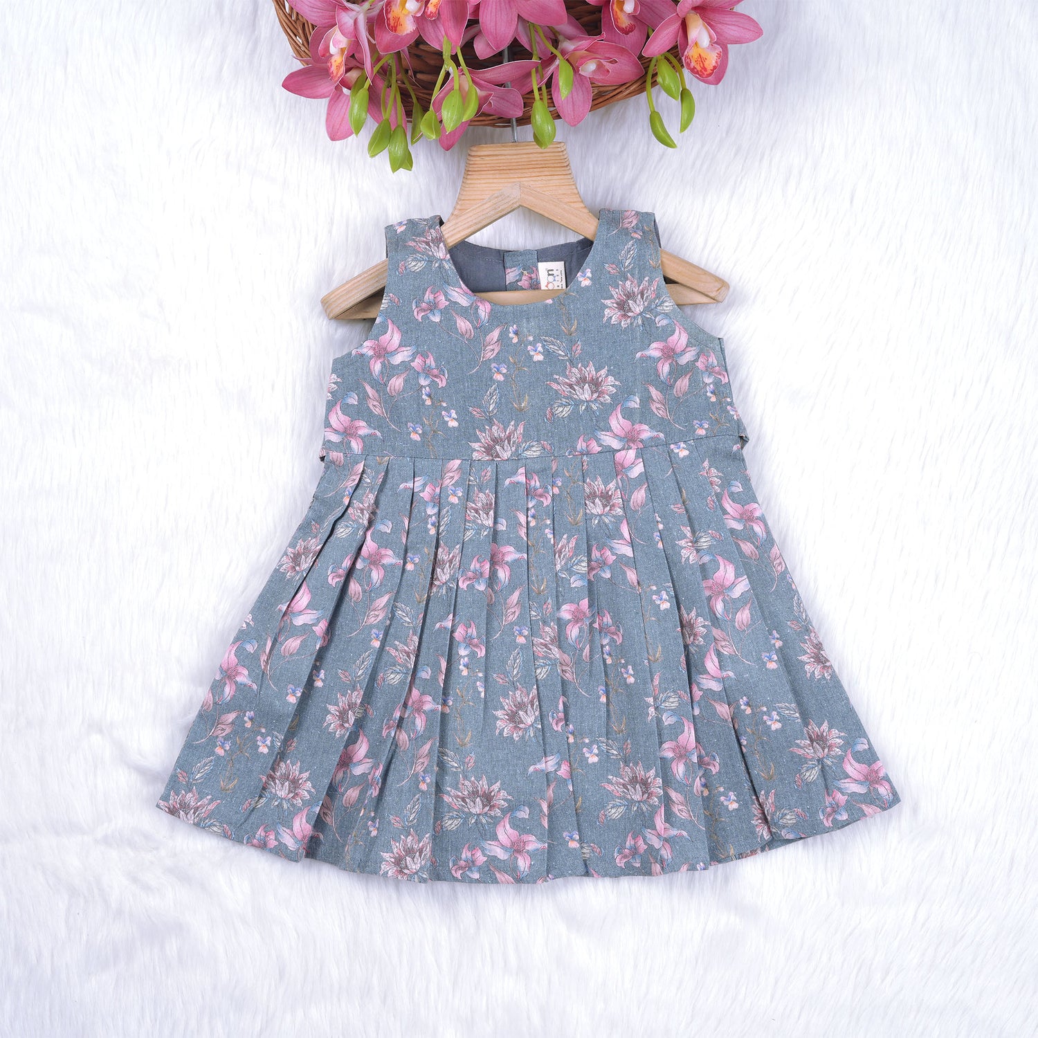 Sleeveless grey cotton frock with pink floral prints, neatly pleated and displayed on a wooden hanger under fresh orchids – from Boon Babies girlswear collection.
