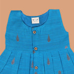 Blue silk sleeveless ethnic frock for baby girls with wooden buttons and traditional motifs, laid flat on a beige background with rustic and floral decor.