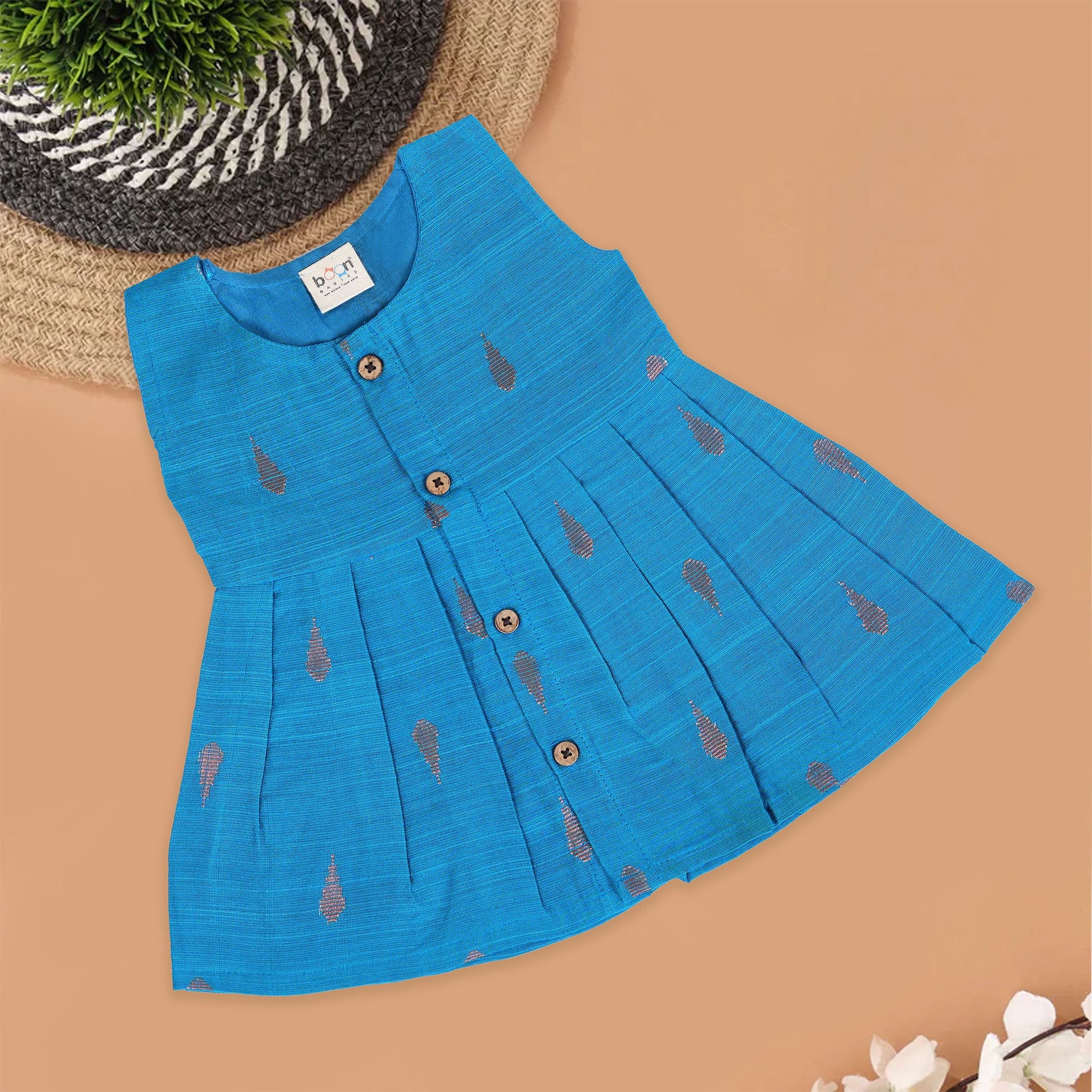 Blue silk sleeveless ethnic frock for baby girls with wooden buttons and traditional motifs, laid flat on a beige background with rustic and floral decor.
