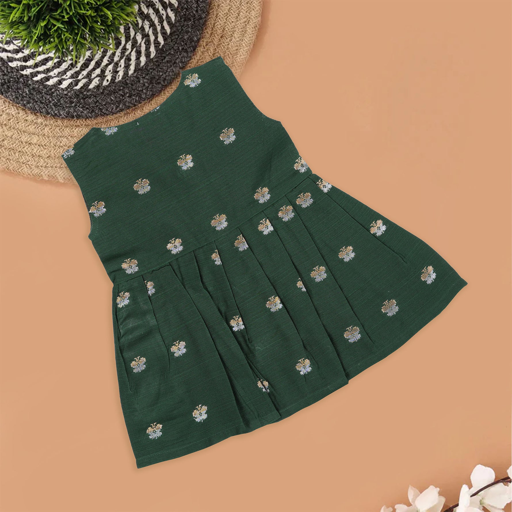 Floral green silk sleeveless frock for baby girls with wooden buttons and subtle motifs, displayed on a beige backdrop with soft floral accents and woven mat.