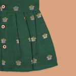 Floral green silk sleeveless frock for baby girls with wooden buttons and subtle motifs, displayed on a beige backdrop with soft floral accents and woven mat.