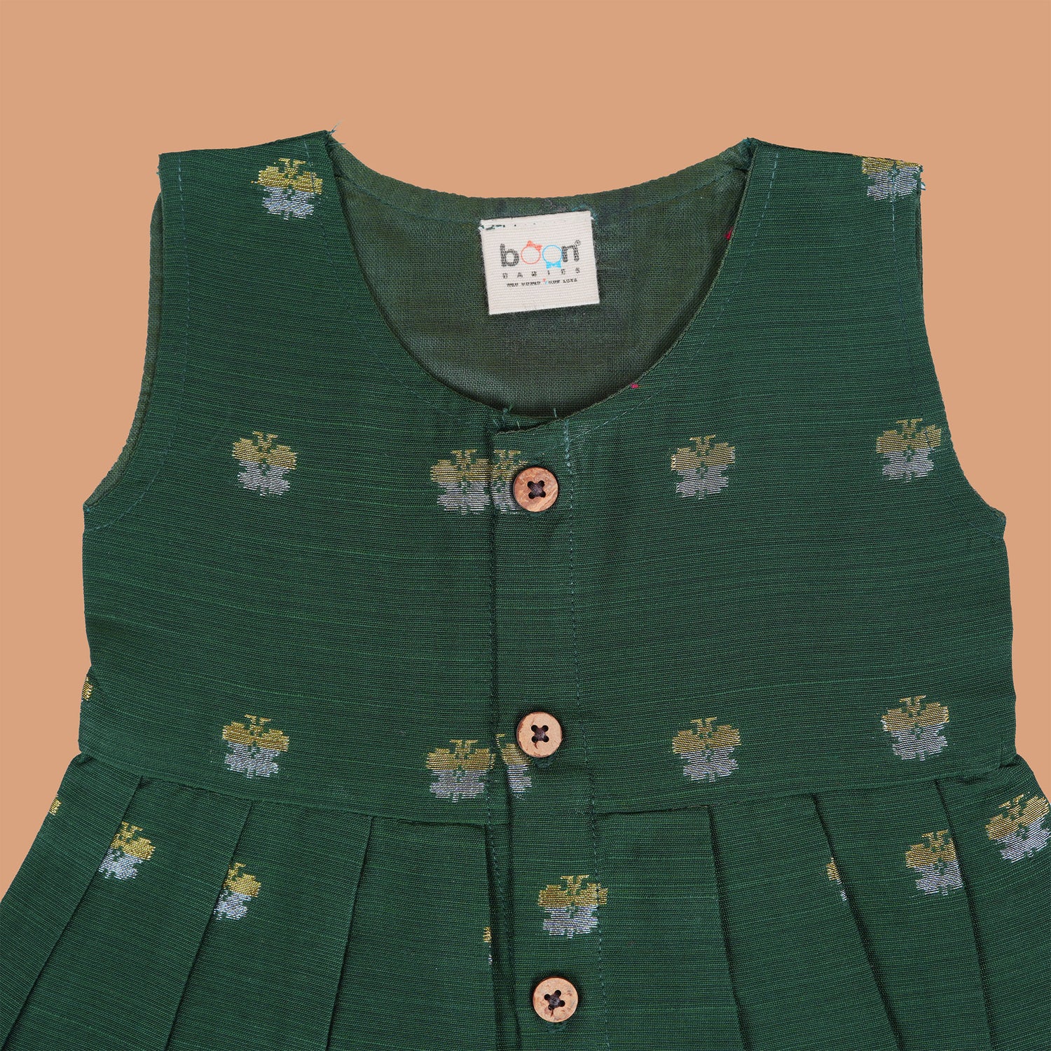 Floral green silk sleeveless frock for baby girls with wooden buttons and subtle motifs, displayed on a beige backdrop with soft floral accents and woven mat.