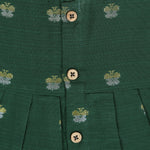 Floral green silk sleeveless frock for baby girls with wooden buttons and subtle motifs, displayed on a beige backdrop with soft floral accents and woven mat.