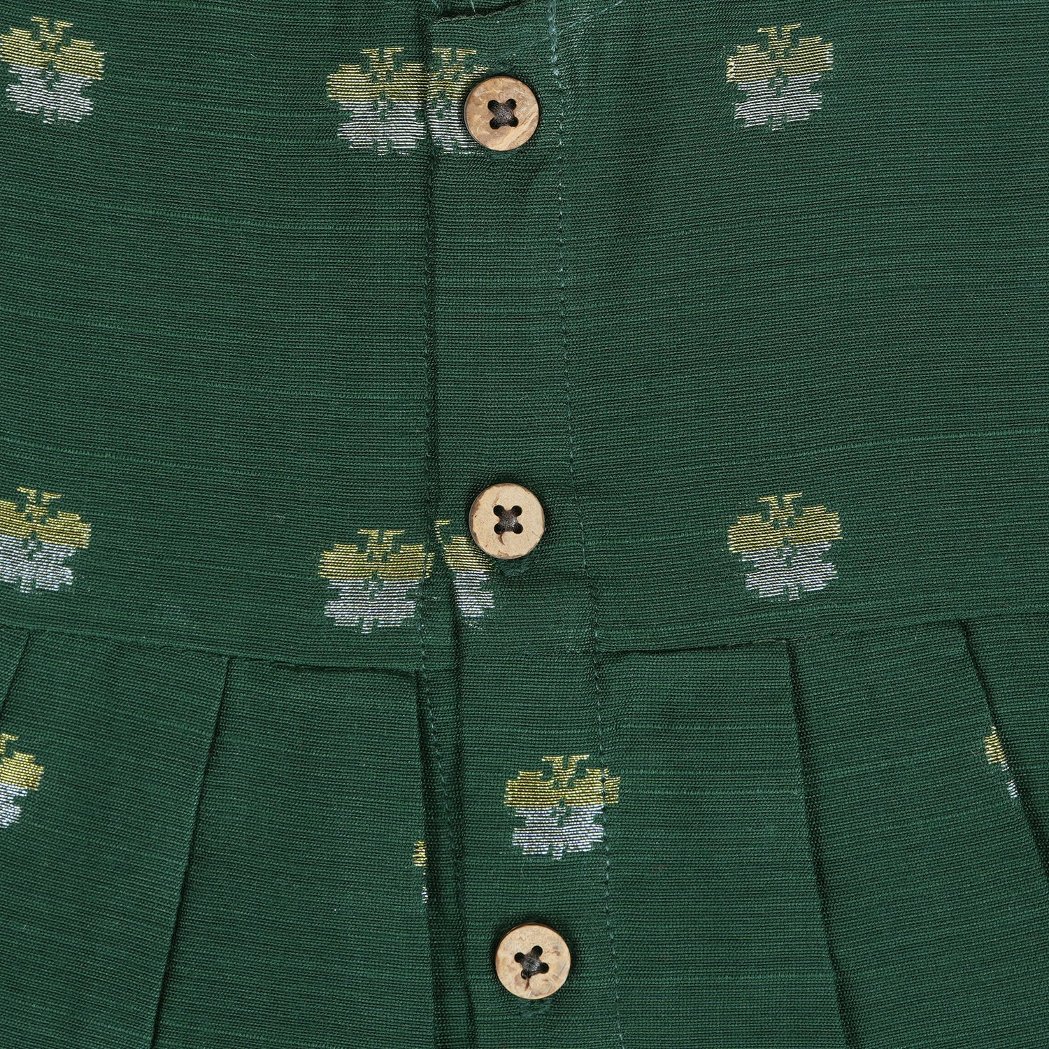 Floral green silk sleeveless frock for baby girls with wooden buttons and subtle motifs, displayed on a beige backdrop with soft floral accents and woven mat.