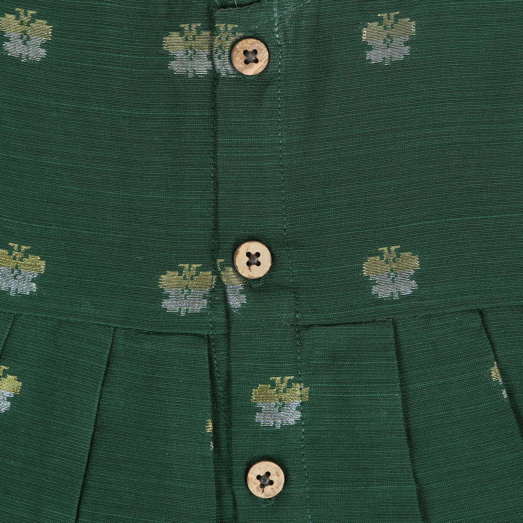 Floral green silk sleeveless frock for baby girls with wooden buttons and subtle motifs, displayed on a beige backdrop with soft floral accents and woven mat.
