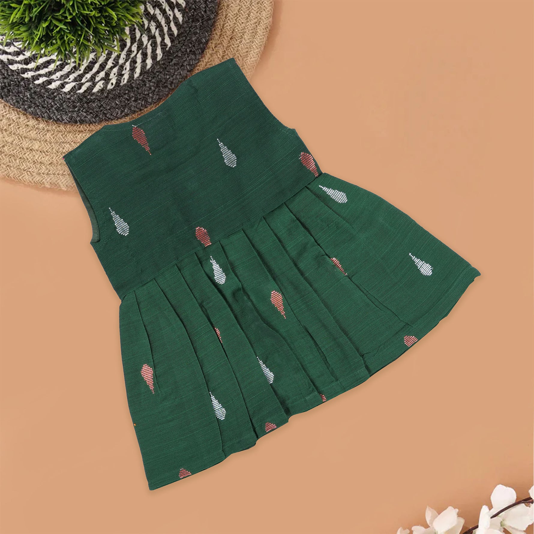 Green ethnic sleeveless silk frock for baby girls with wooden buttons and ikat-style motifs, laid flat on a beige background with natural decor and floral accents.