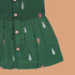 Green ethnic sleeveless silk frock for baby girls with wooden buttons and ikat-style motifs, laid flat on a beige background with natural decor and floral accents.