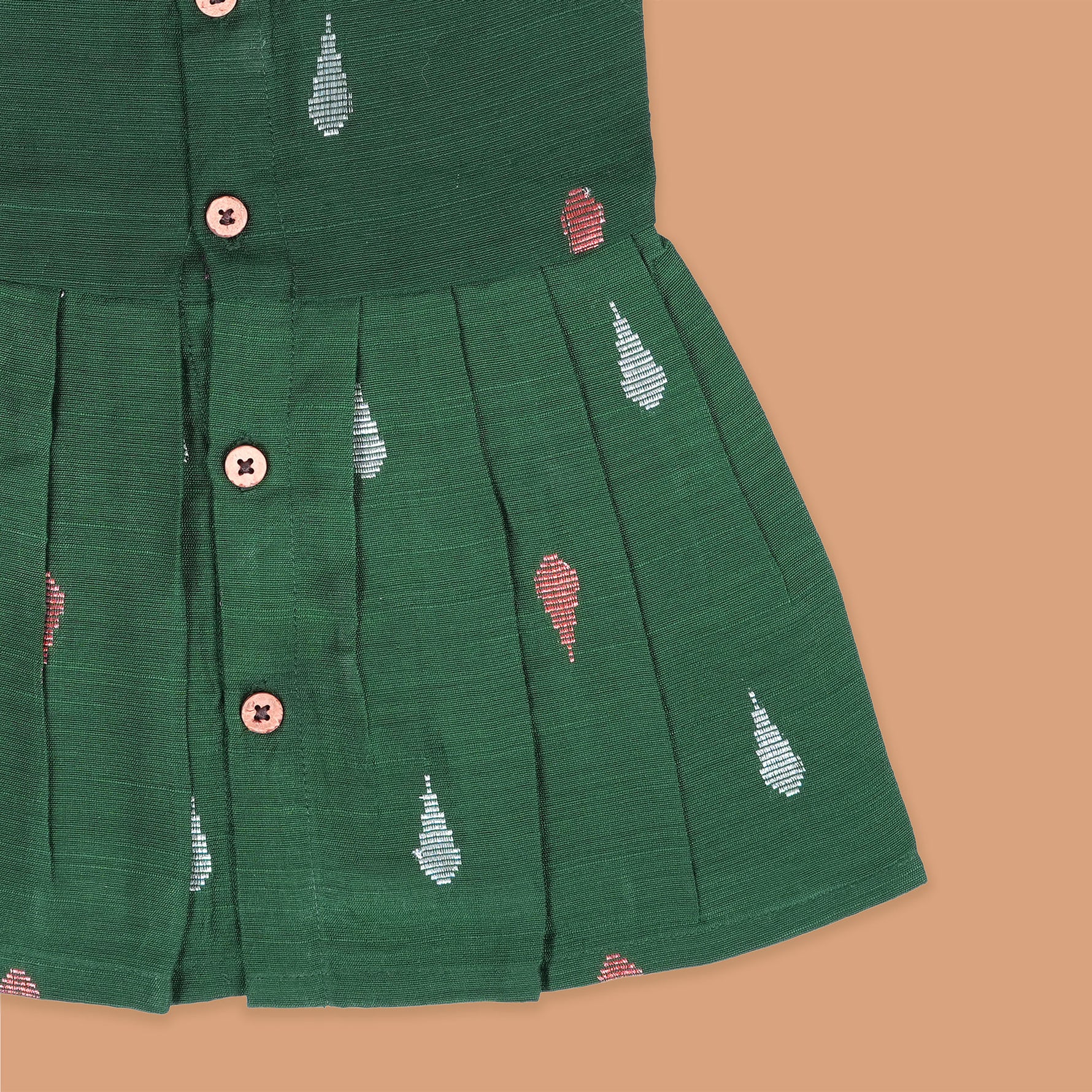 Green ethnic sleeveless silk frock for baby girls with wooden buttons and ikat-style motifs, laid flat on a beige background with natural decor and floral accents.