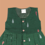 Green ethnic sleeveless silk frock for baby girls with wooden buttons and ikat-style motifs, laid flat on a beige background with natural decor and floral accents.