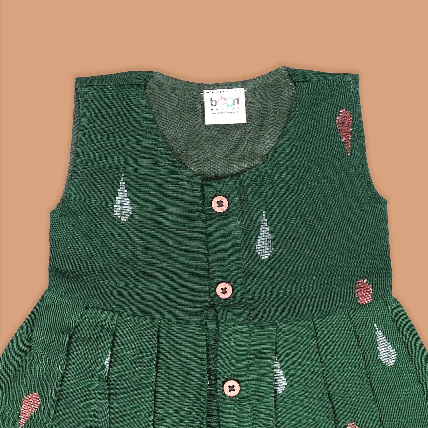 Green ethnic sleeveless silk frock for baby girls with wooden buttons and ikat-style motifs, laid flat on a beige background with natural decor and floral accents.