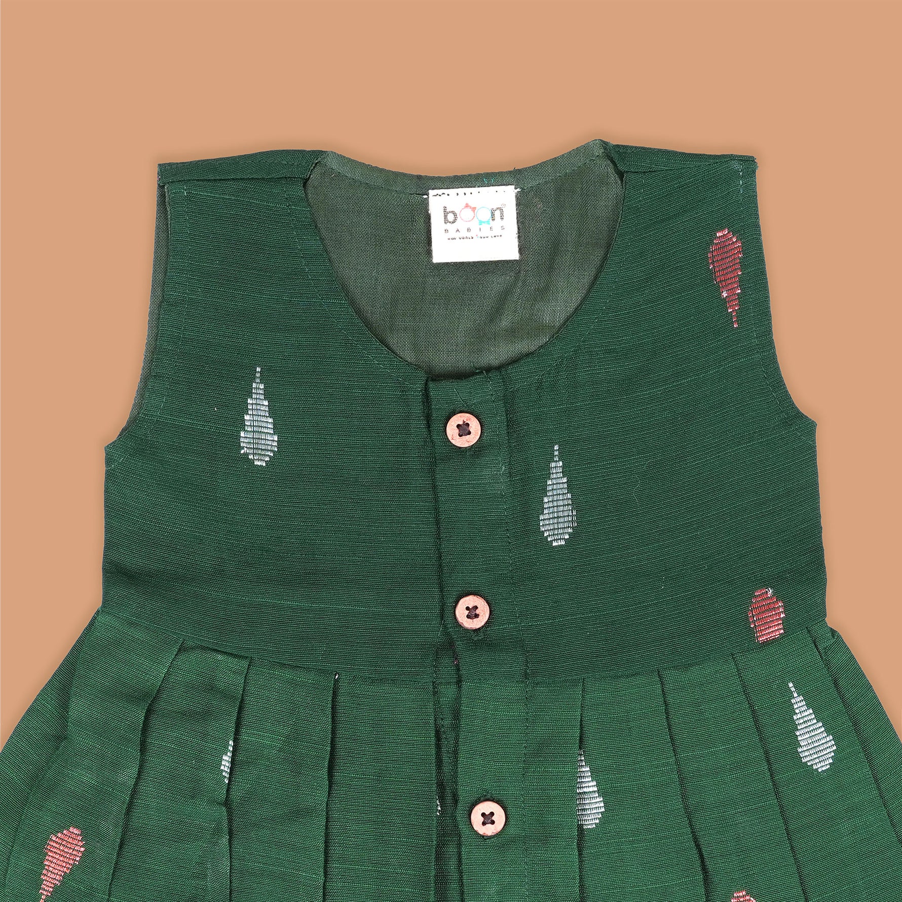 Green ethnic sleeveless silk frock for baby girls with wooden buttons and ikat-style motifs, laid flat on a beige background with natural decor and floral accents.