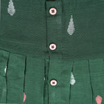 Green ethnic sleeveless silk frock for baby girls with wooden buttons and ikat-style motifs, laid flat on a beige background with natural decor and floral accents.