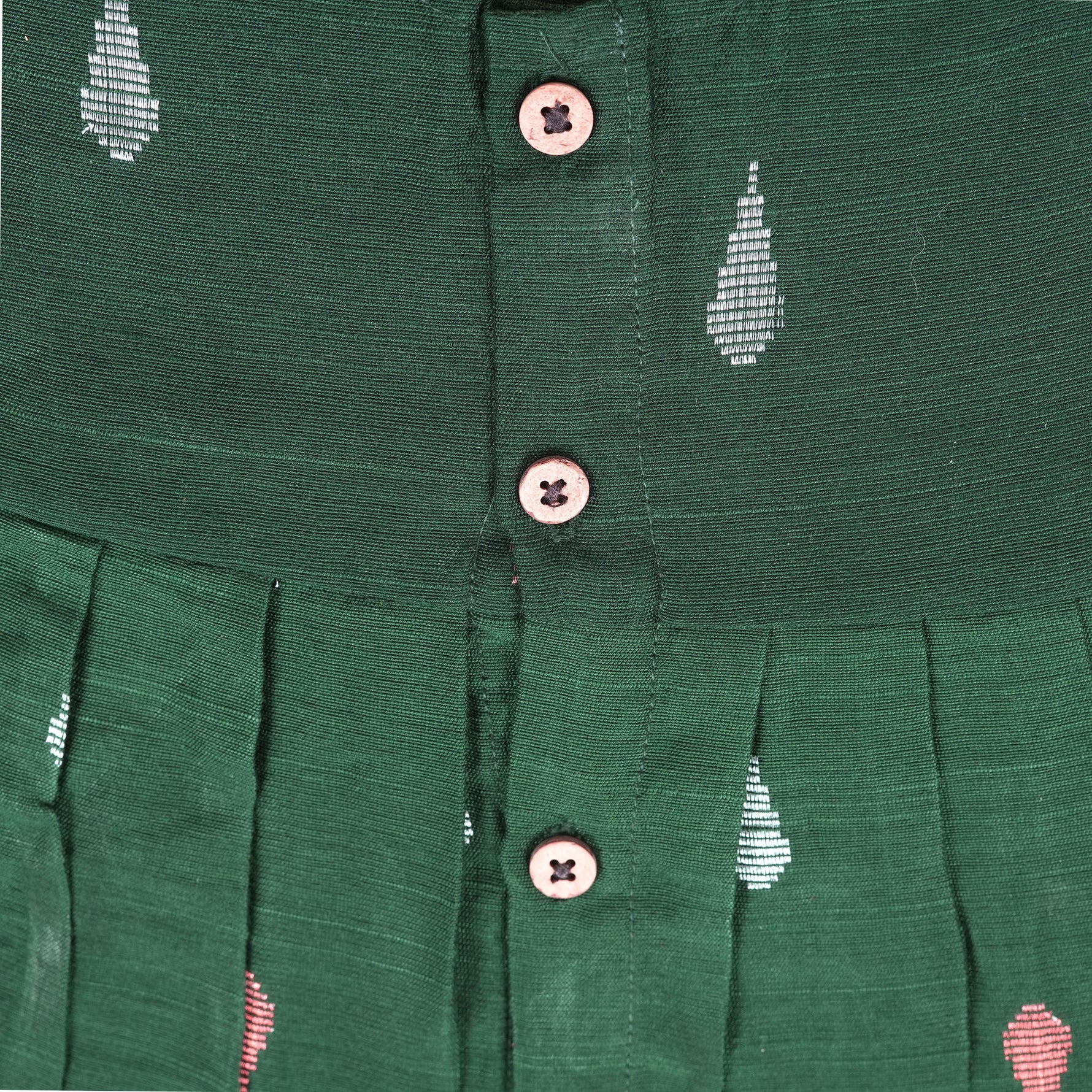 Green ethnic sleeveless silk frock for baby girls with wooden buttons and ikat-style motifs, laid flat on a beige background with natural decor and floral accents.