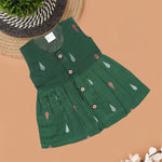 Green ethnic sleeveless silk frock for baby girls with wooden buttons and ikat-style motifs, laid flat on a beige background with natural decor and floral accents.