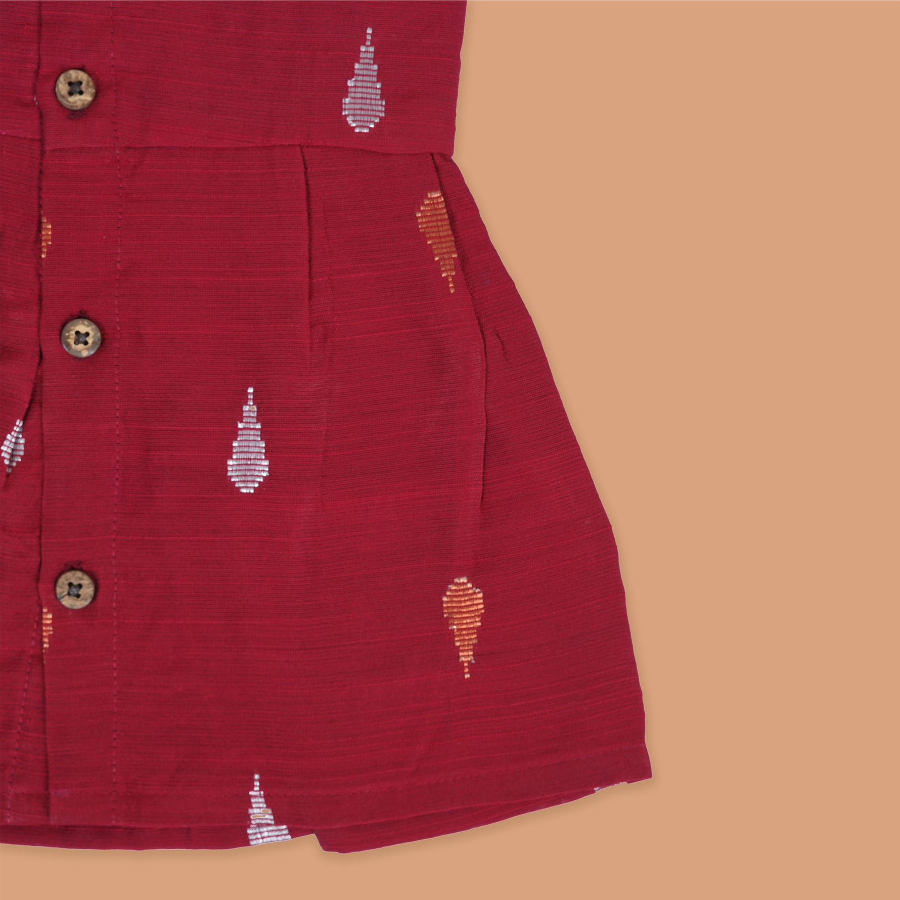 Maroon sleeveless ethnic silk frock for baby girls with wooden buttons and traditional motifs, displayed on a beige background with rustic mats and floral accents.