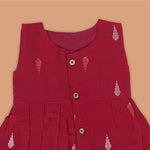 Maroon sleeveless ethnic silk frock for baby girls with wooden buttons and traditional motifs, displayed on a beige background with rustic mats and floral accents.