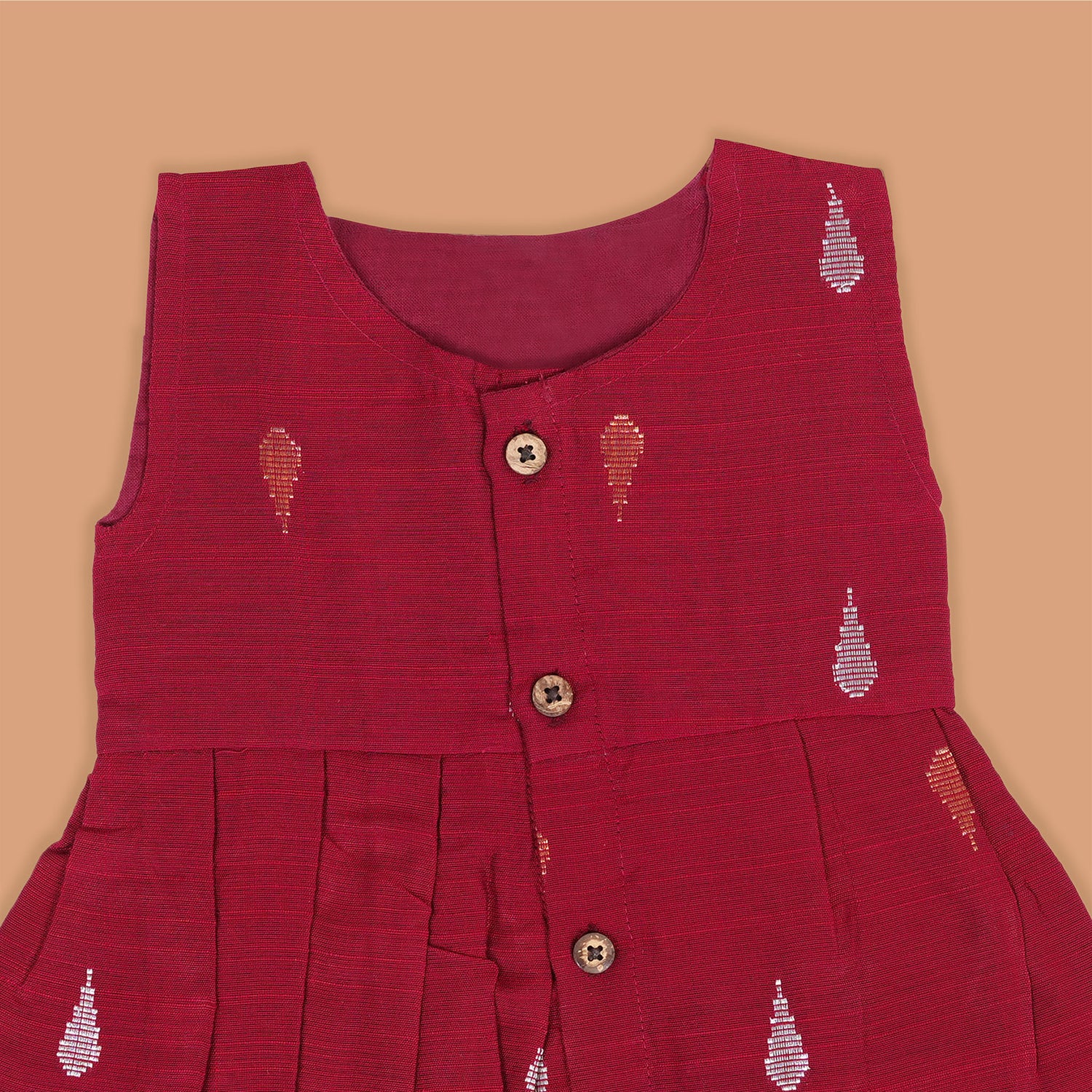 Maroon sleeveless ethnic silk frock for baby girls with wooden buttons and traditional motifs, displayed on a beige background with rustic mats and floral accents.