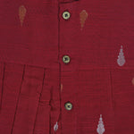 Maroon sleeveless ethnic silk frock for baby girls with wooden buttons and traditional motifs, displayed on a beige background with rustic mats and floral accents.