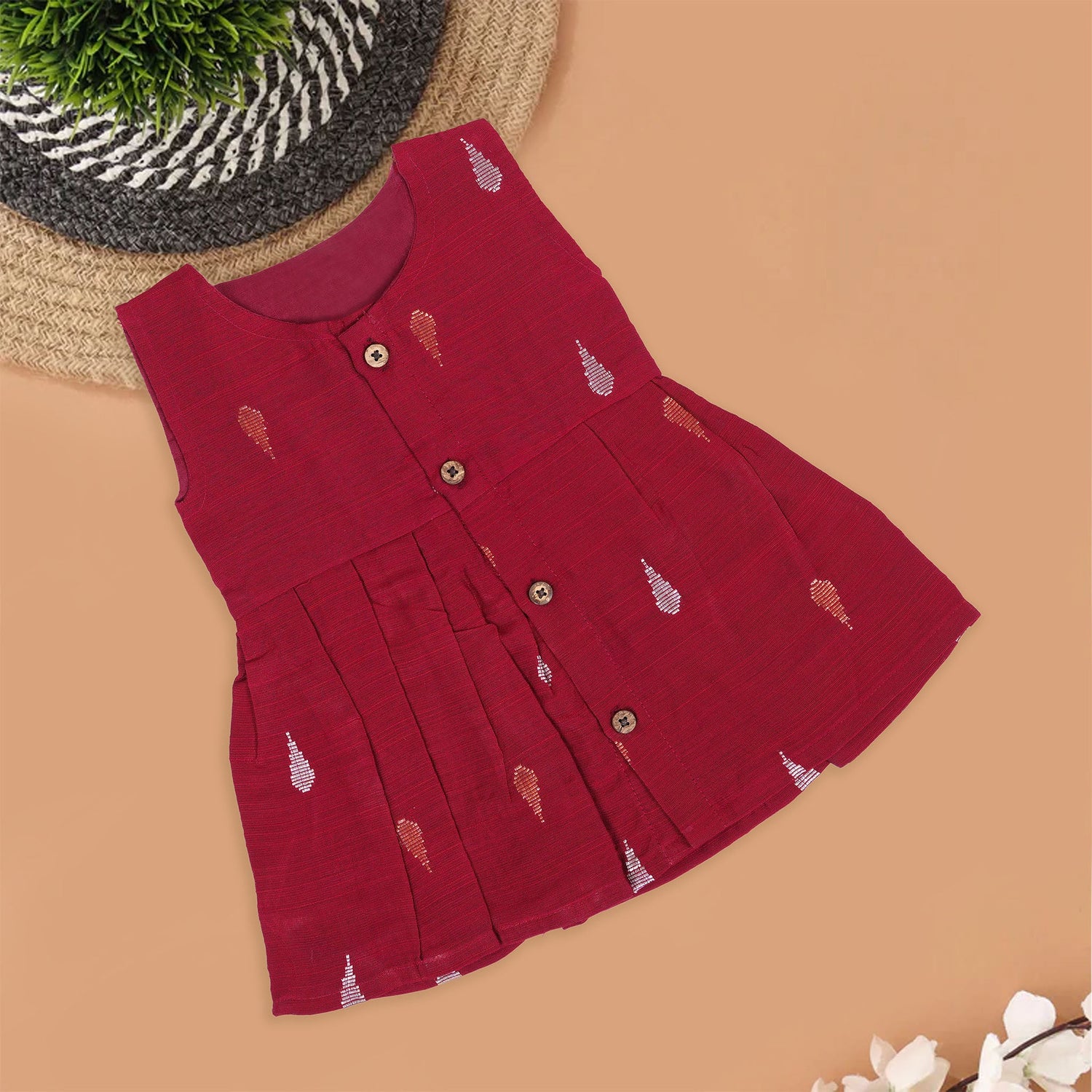 Maroon sleeveless ethnic silk frock for baby girls with wooden buttons and traditional motifs, displayed on a beige background with rustic mats and floral accents.