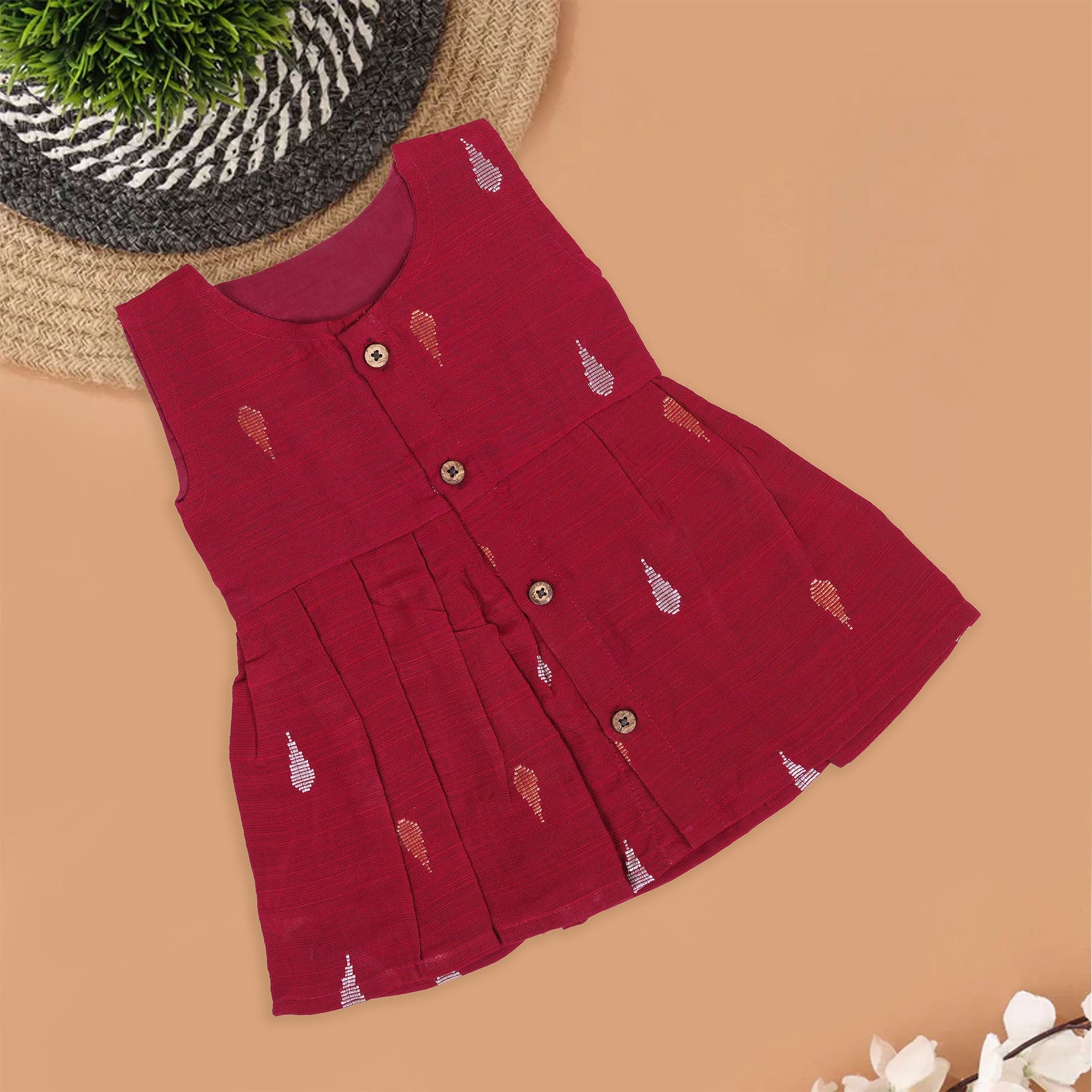 Maroon sleeveless ethnic silk frock for baby girls with wooden buttons and traditional motifs, displayed on a beige background with rustic mats and floral accents.