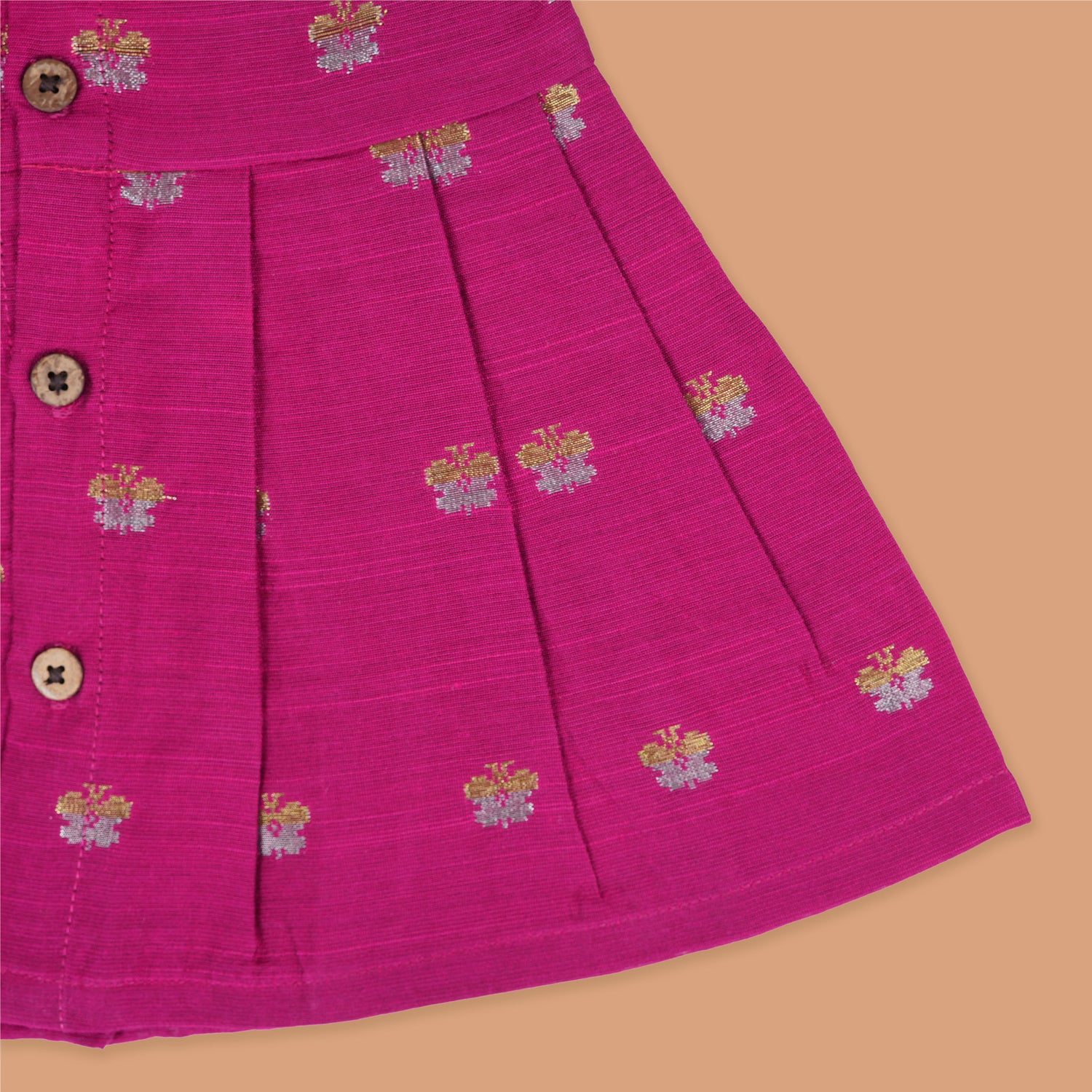 Pink silk sleeveless ethnic frock for baby girls with woven floral motifs and wooden buttons, displayed flat on a peach background with natural and floral props.