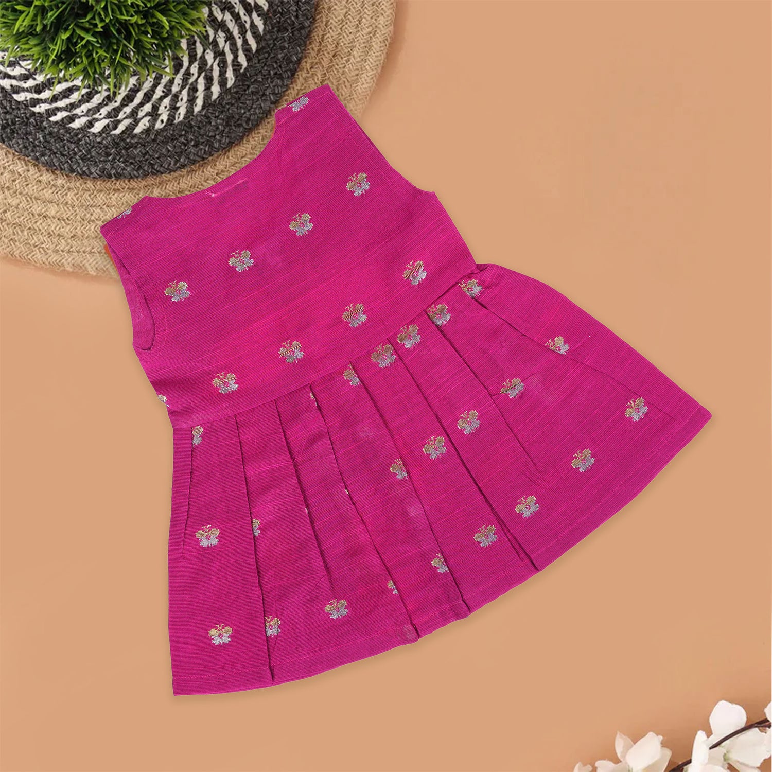 Pink silk sleeveless ethnic frock for baby girls with woven floral motifs and wooden buttons, displayed flat on a peach background with natural and floral props.