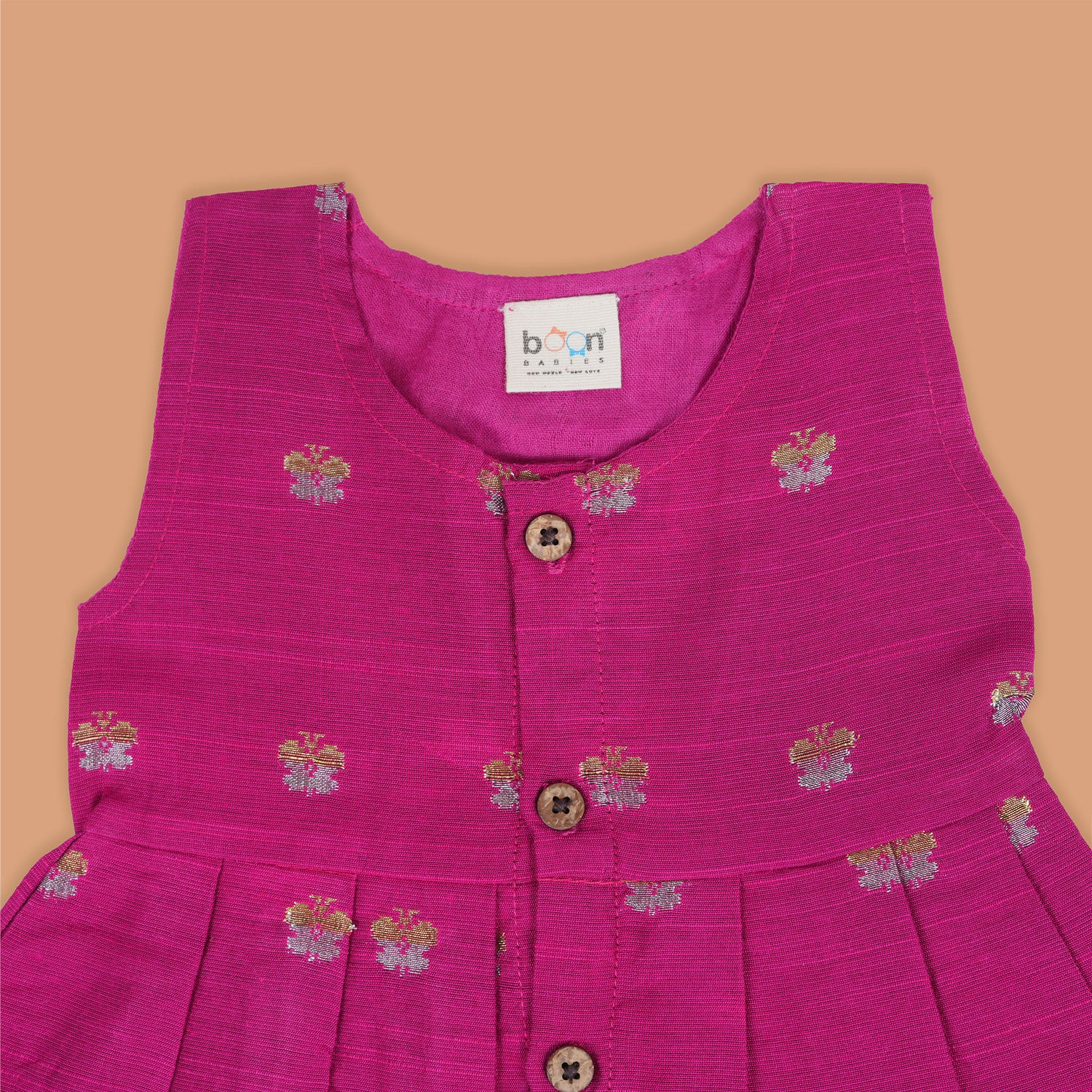 Pink silk sleeveless ethnic frock for baby girls with woven floral motifs and wooden buttons, displayed flat on a peach background with natural and floral props.