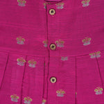 Pink silk sleeveless ethnic frock for baby girls with woven floral motifs and wooden buttons, displayed flat on a peach background with natural and floral props.