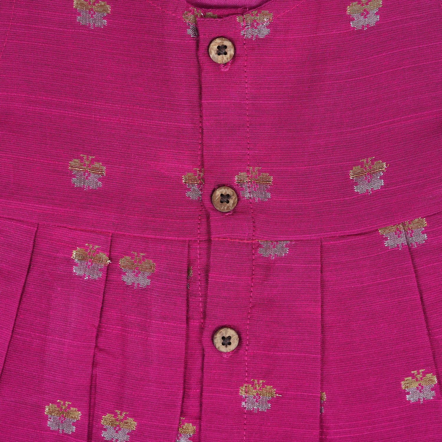 Pink silk sleeveless ethnic frock for baby girls with woven floral motifs and wooden buttons, displayed flat on a peach background with natural and floral props.