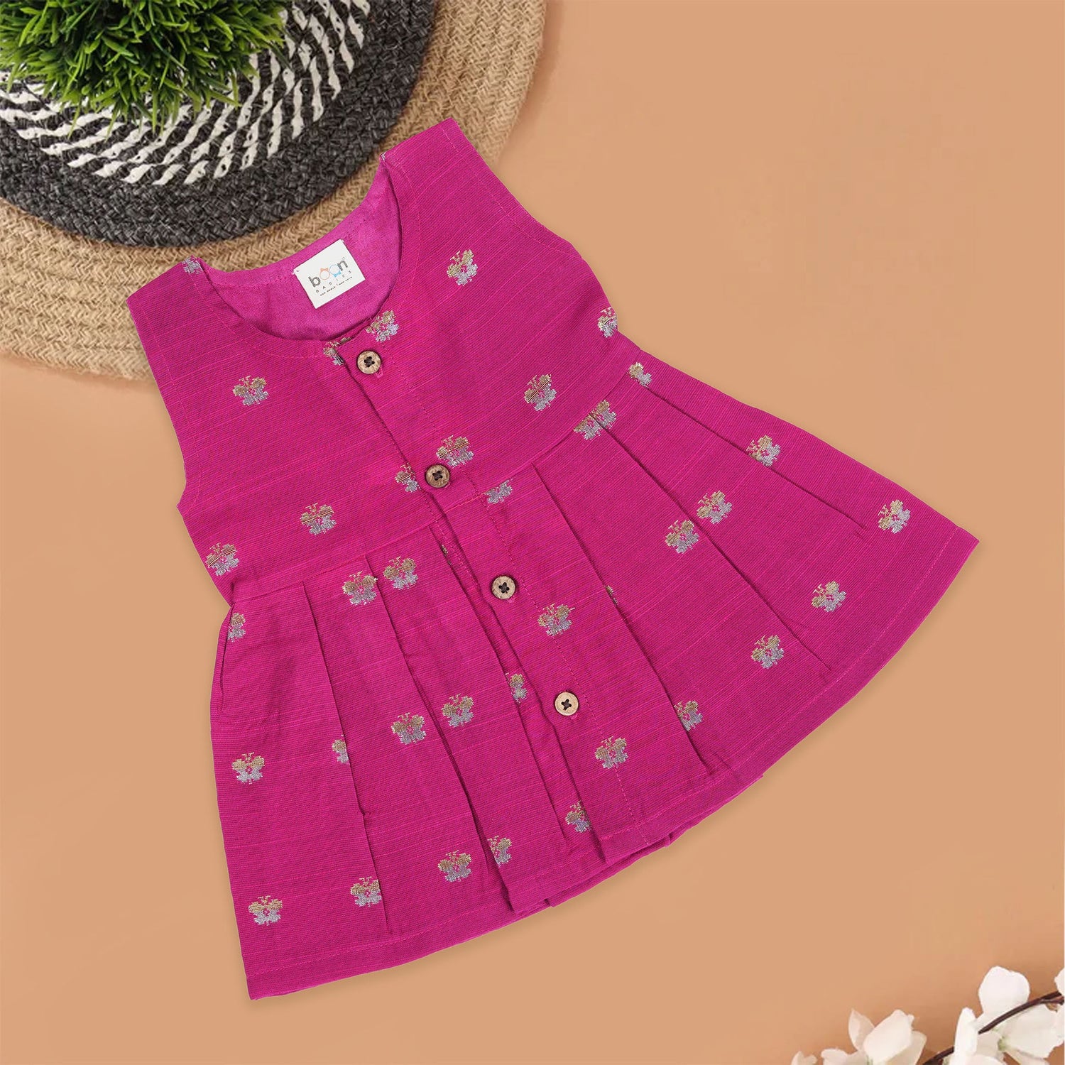 Pink silk sleeveless ethnic frock for baby girls with woven floral motifs and wooden buttons, displayed flat on a peach background with natural and floral props.
