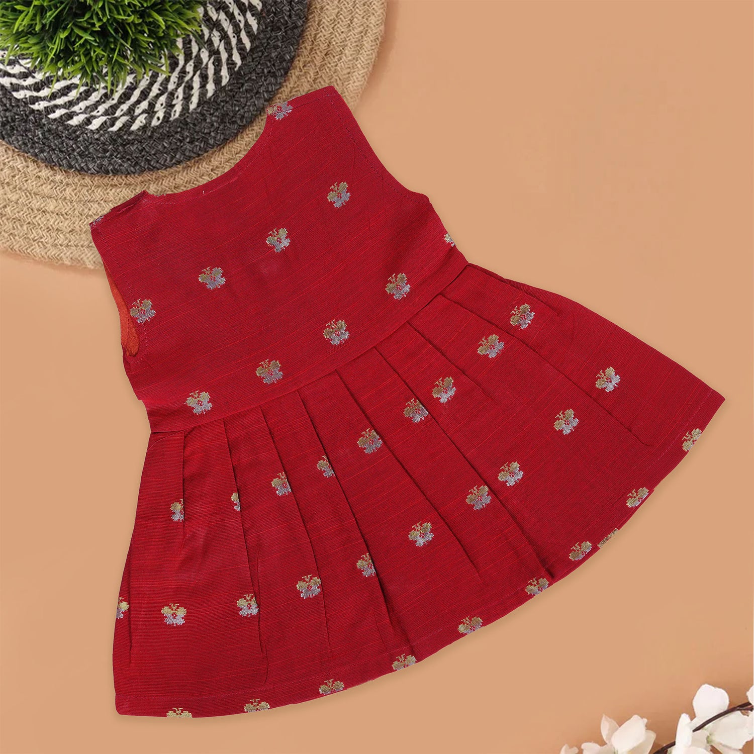 Red silk sleeveless ethnic frock for baby girls with floral woven motifs and wooden button closure, laid flat on a beige background with white florals and rustic decor.