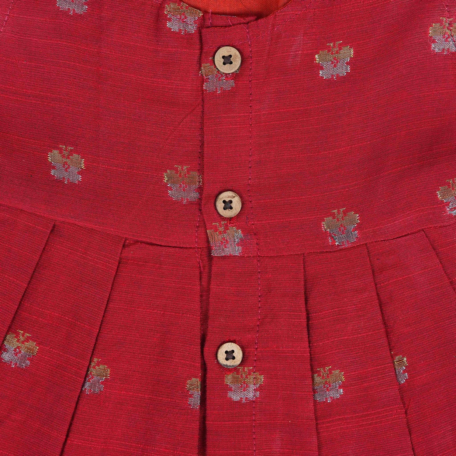 Red silk sleeveless ethnic frock for baby girls with floral woven motifs and wooden button closure, laid flat on a beige background with white florals and rustic decor.
