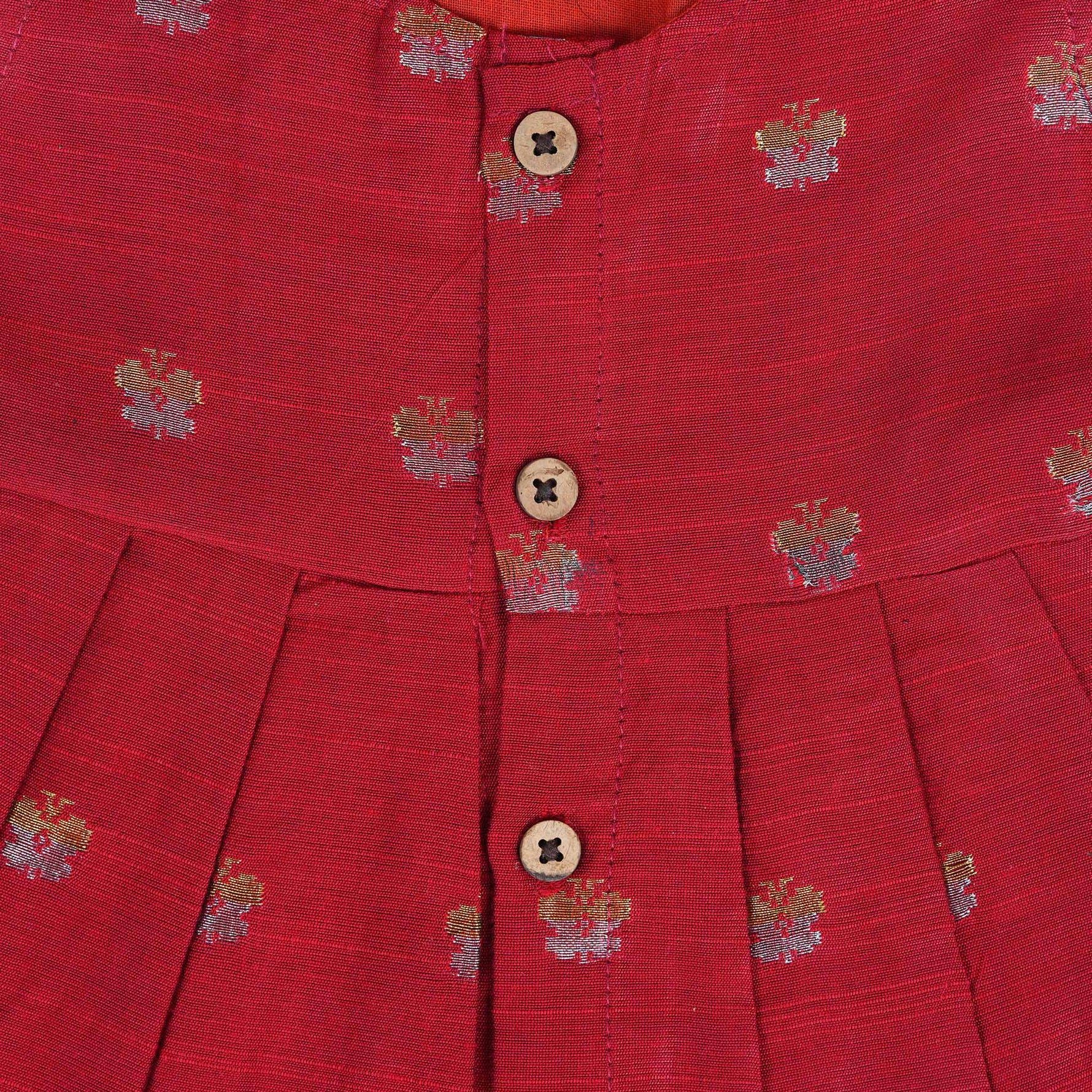 Red silk sleeveless ethnic frock for baby girls with floral woven motifs and wooden button closure, laid flat on a beige background with white florals and rustic decor.