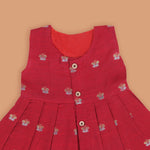 Red silk sleeveless ethnic frock for baby girls with floral woven motifs and wooden button closure, laid flat on a beige background with white florals and rustic decor.