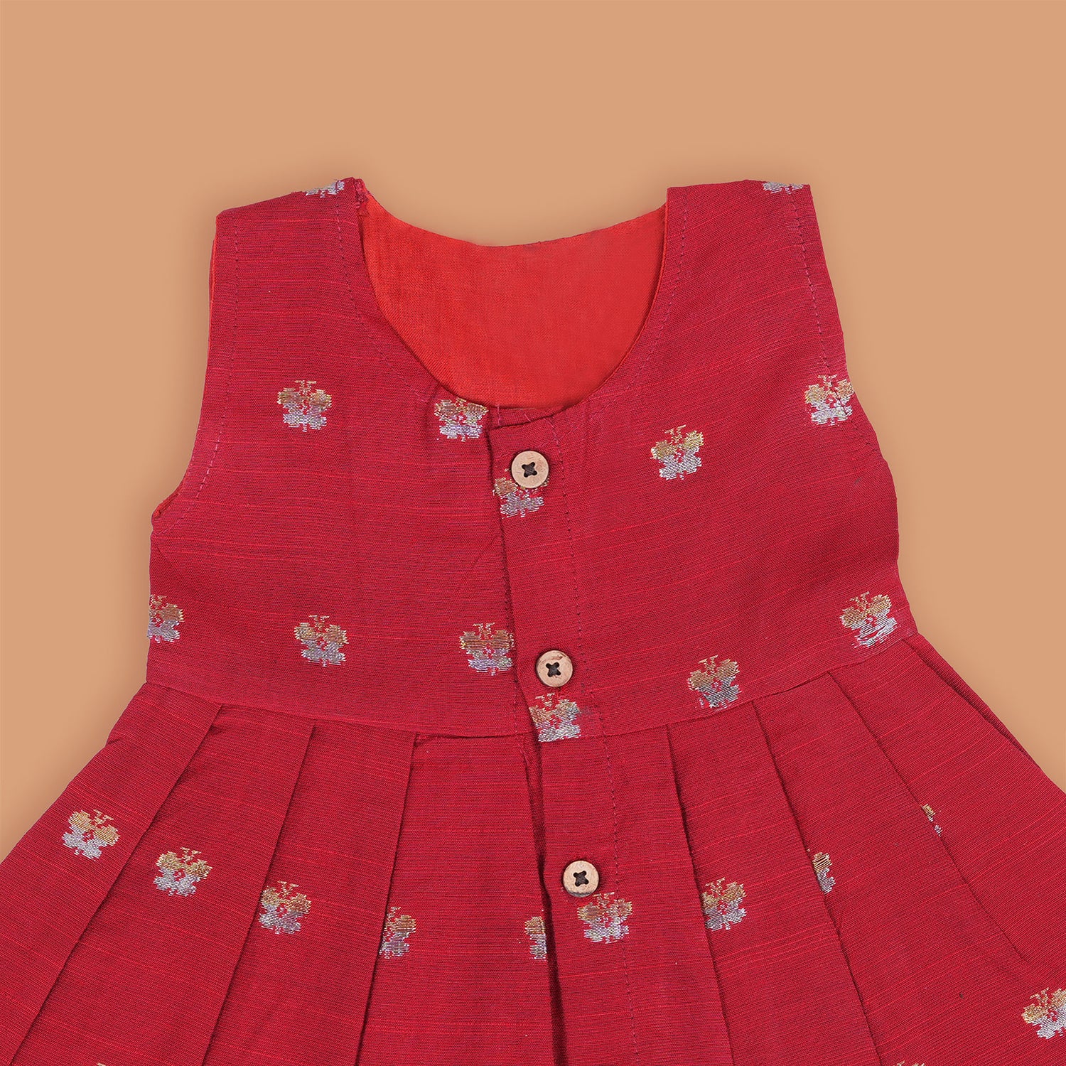 Red silk sleeveless ethnic frock for baby girls with floral woven motifs and wooden button closure, laid flat on a beige background with white florals and rustic decor.