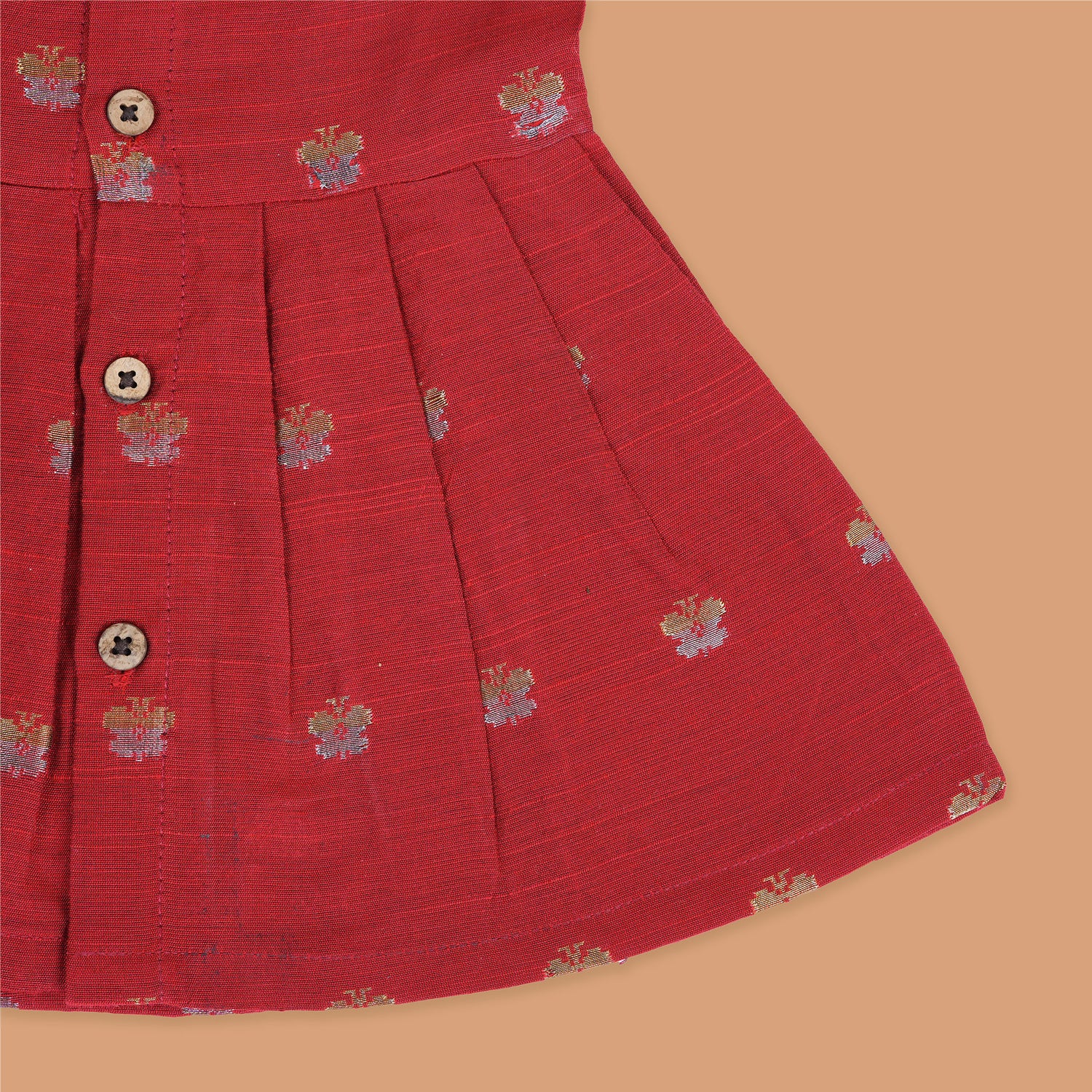Red silk sleeveless ethnic frock for baby girls with floral woven motifs and wooden button closure, laid flat on a beige background with white florals and rustic decor.