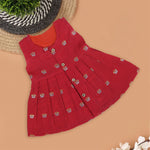 Red silk sleeveless ethnic frock for baby girls with floral woven motifs and wooden button closure, laid flat on a beige background with white florals and rustic decor.